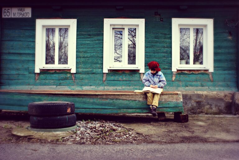 Child reading a book outside a rustic green house, under a street sign, conveying solitude and curiosity.
